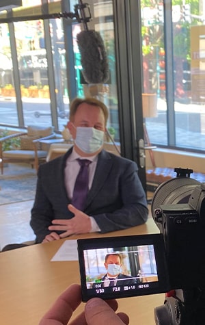 Man in suit and surgical mask being interviewed on video camera with boom mic visible overhead