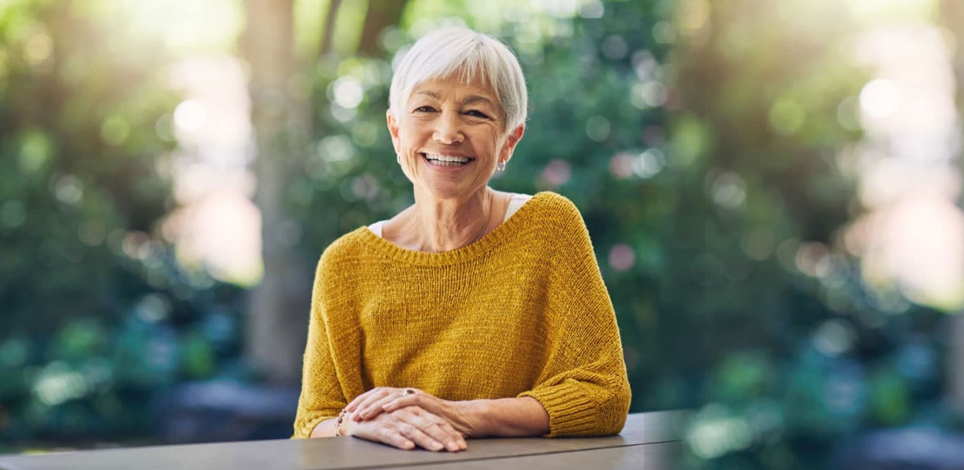 Elderly woman in yellow sweater leaning on fence outside and smiling.